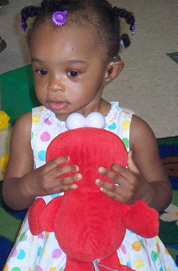 A toddler with a hearing aid holds a red stuffed animal